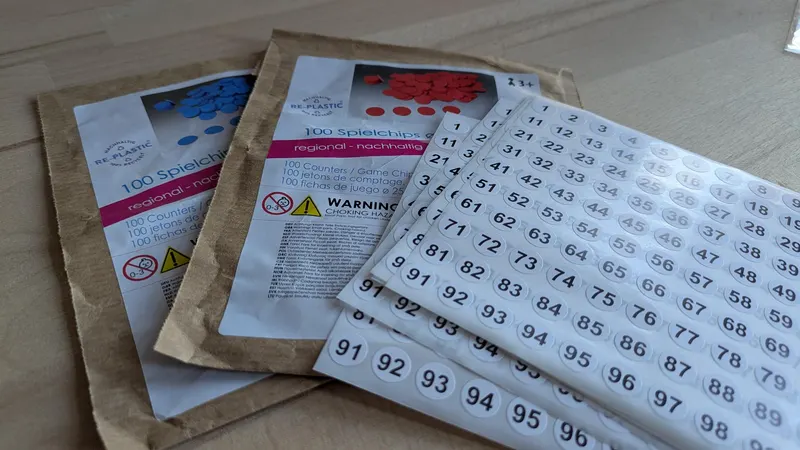 Zwei Tüten mit je 100 Spielchips in Blau und Rot, daneben Bögen mit runden Zahlenaufklebern